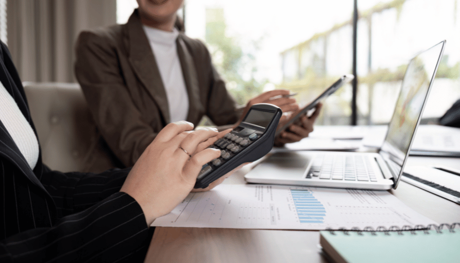 People in business attire using a calculator and laptop during a meeting at a desk.