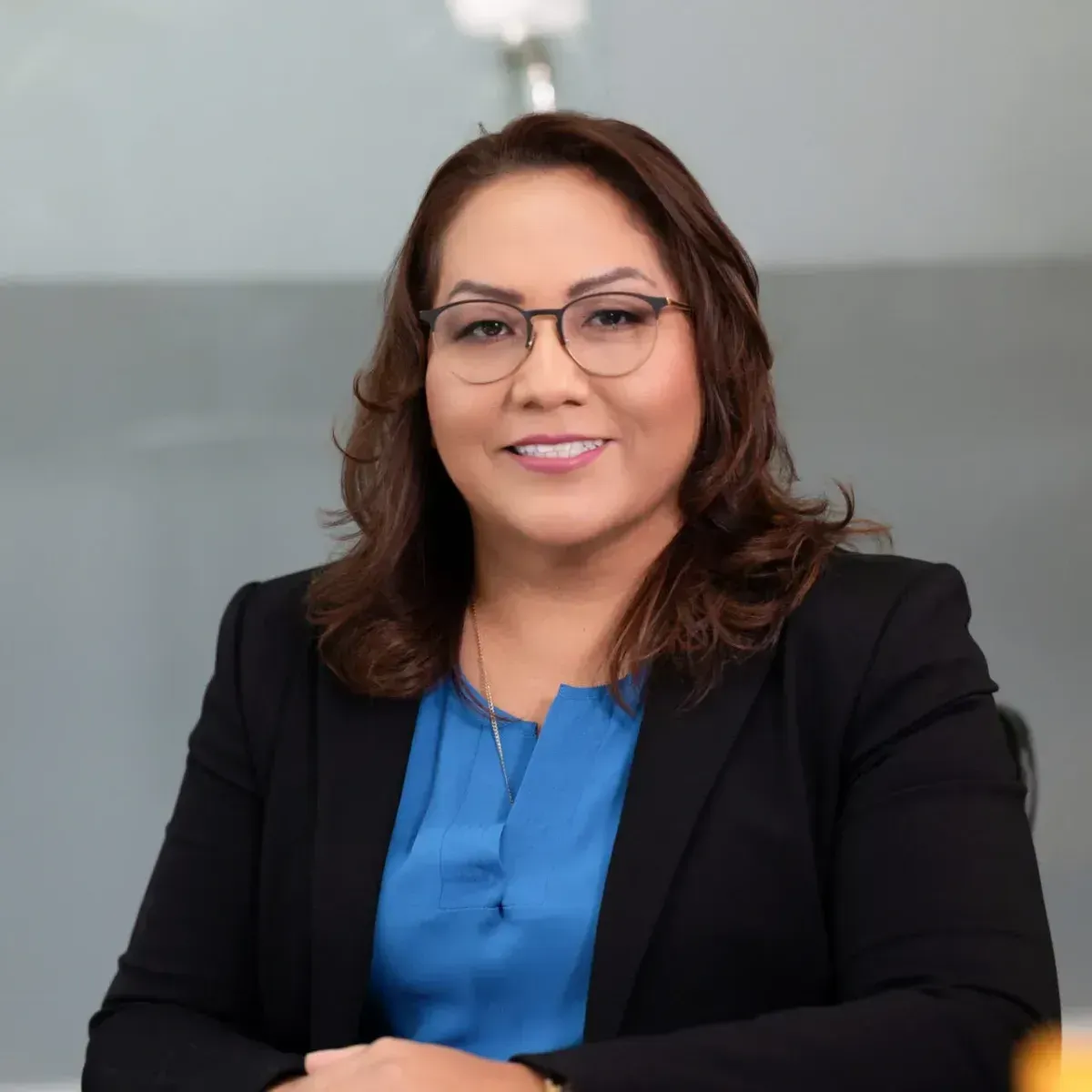 Woman in black blazer and blue blouse, smiling with arms crossed against a gray background