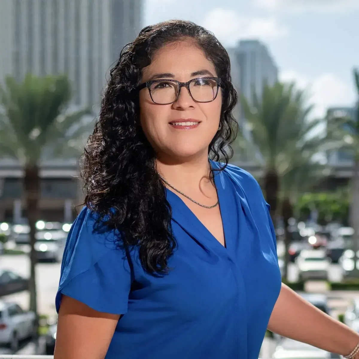 Woman in blue blouse smiling outdoors with city buildings and palm trees in the background