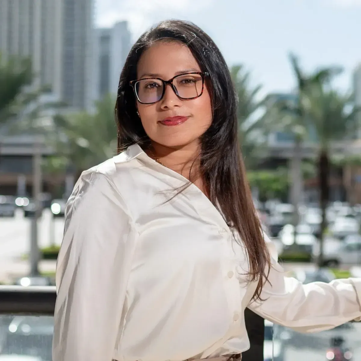 Woman in white blouse and glasses standing outdoors with palm trees and city buildings in the background