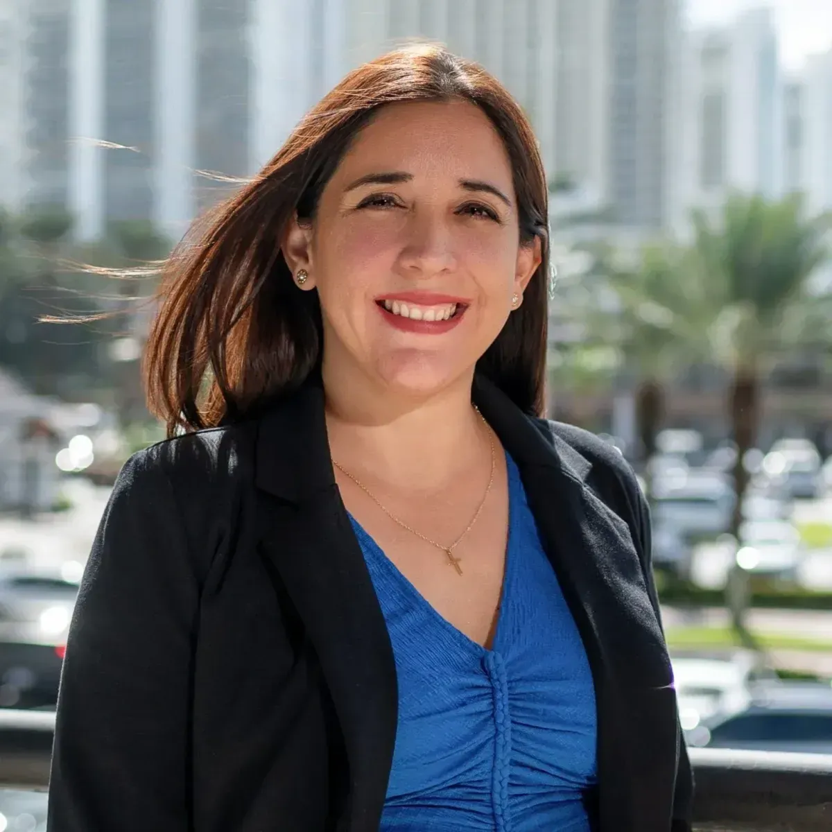 Smiling woman in a black blazer and blue top standing outdoors with city buildings behind her