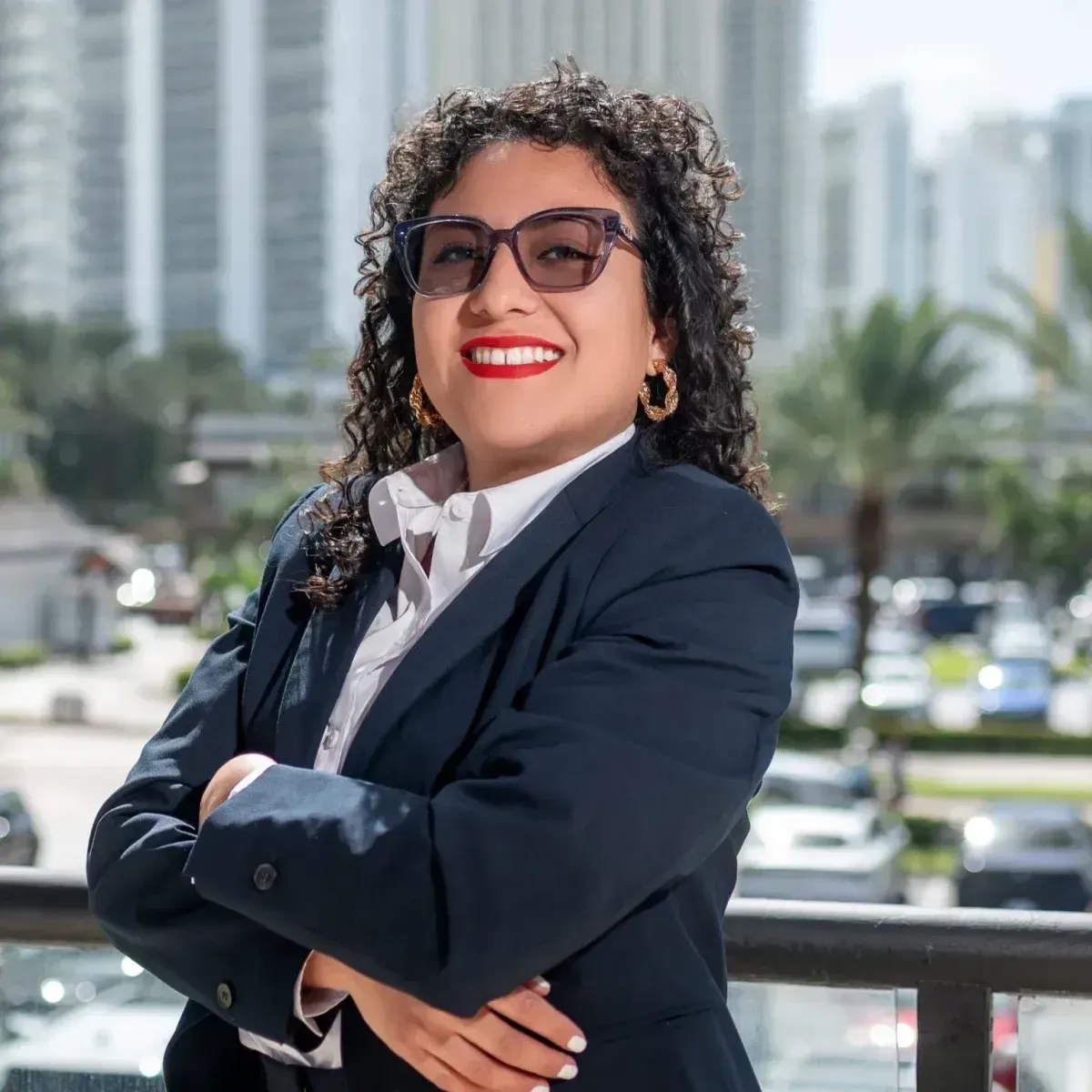 Smiling woman in a black blazer and blue top standing outdoors with city buildings behind her