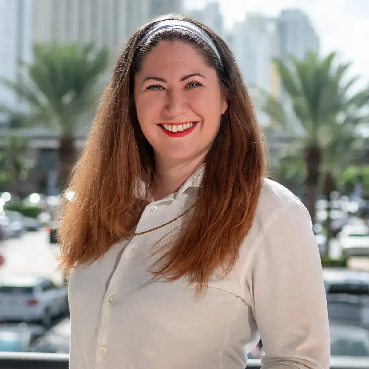 Smiling woman in a white blouse standing outdoors with palm trees and buildings in the background