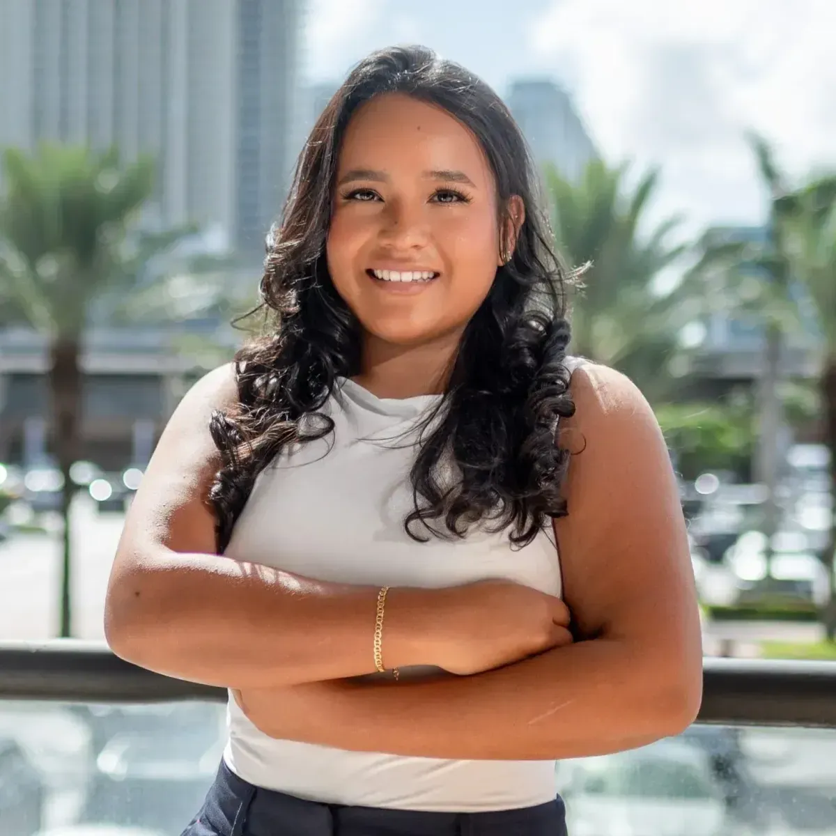 Smiling person with folded arms standing outdoors in front of city buildings and palm trees