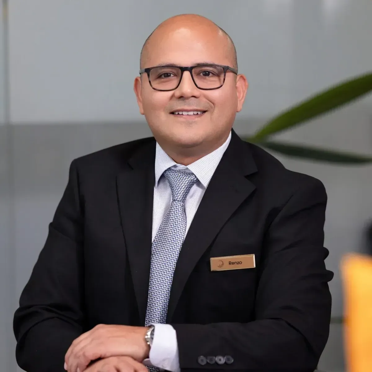 Smiling man in a black suit and glasses seated indoors with a name tag on his jacket