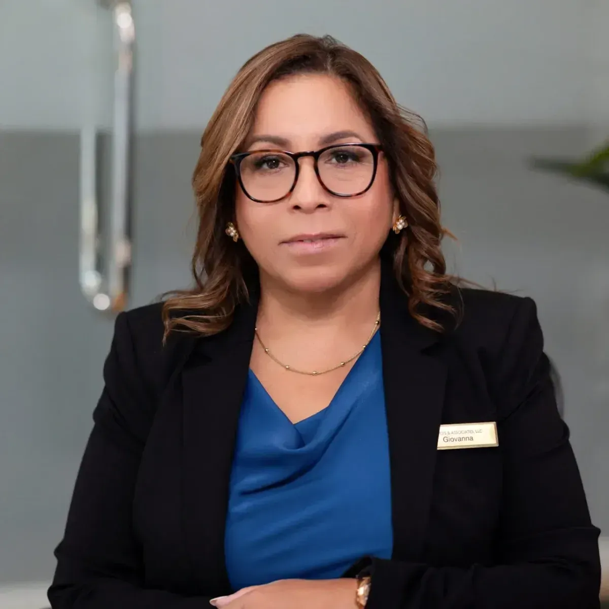 Woman in glasses and black blazer with blue blouse, seated against a gray office background.
