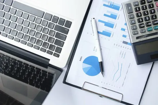Laptop, clipboard with blue charts, pen, and calculator on a desk with paperwork.
