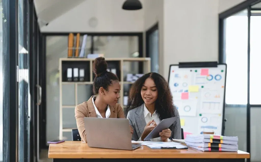 Two coworkers collaborating at a desk with a laptop and notes in a bright office