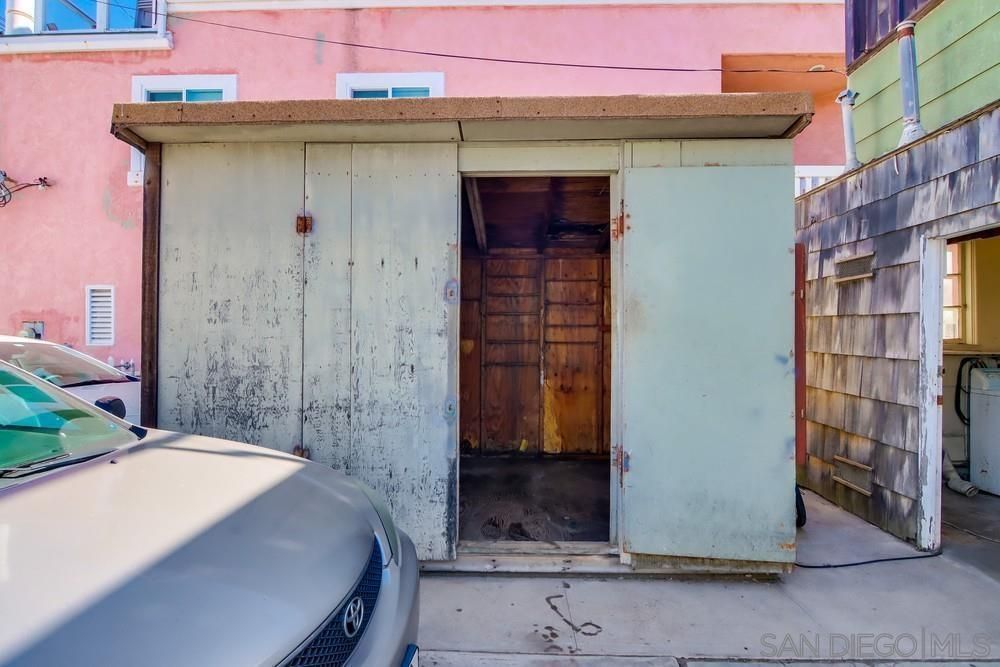A car is parked in front of a shed with a pink building in the background.