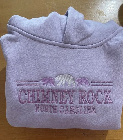 Embroidered hoodie with Chimney Rock Village branding folded on a rustic wooden table.