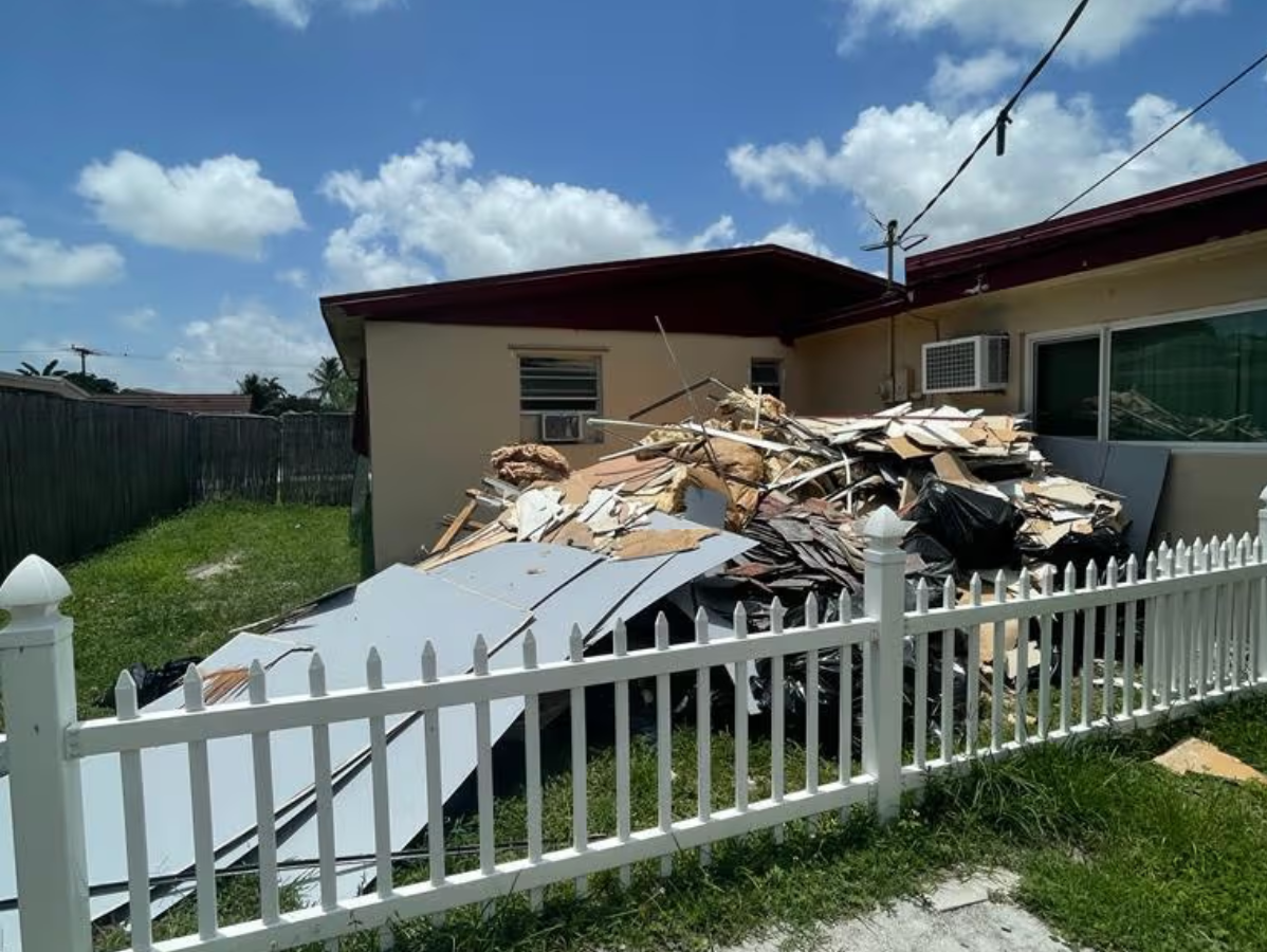 Pile of demolition debris in a yard next to a house with a white picket fence, under a blue sky.