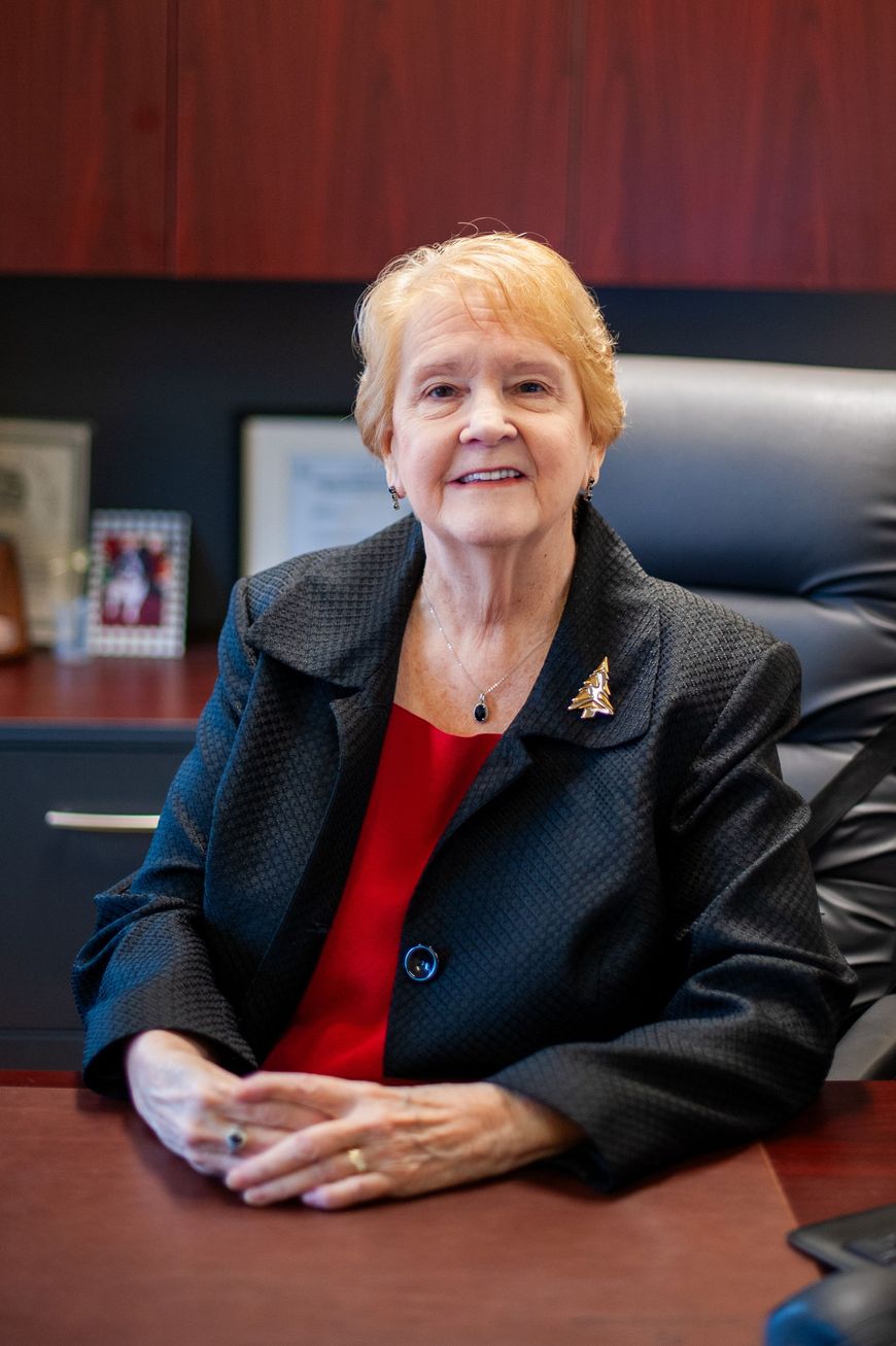 Woman seated at a desk in an office, wearing a dark blazer and red top, smiling at the camera