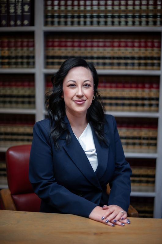 Woman in a navy blazer seated at a desk in front of law books, smiling in an office setting