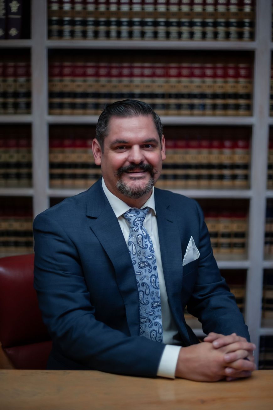 Man in a navy suit seated at a desk, smiling in front of a bookshelf backdrop.