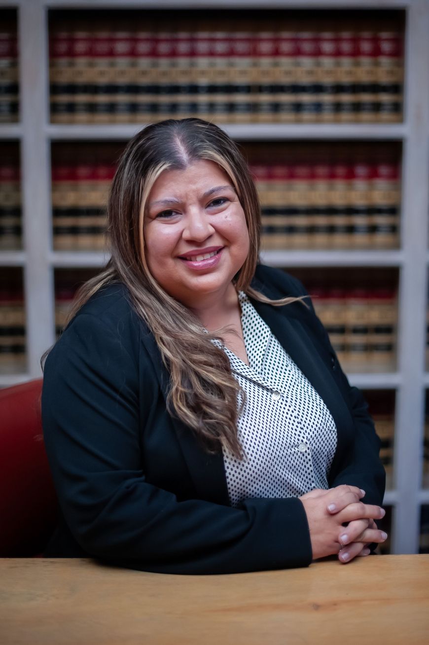 Smiling woman in a black blazer seated at a desk in front of bookshelves with legal books