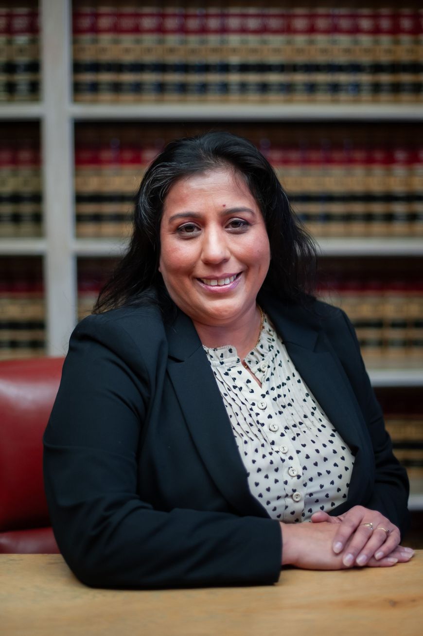 Smiling person in a black blazer seated at a table in front of bookshelves.