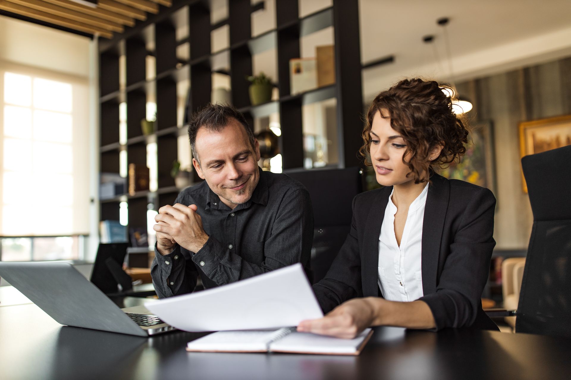 Two coworkers review papers at a table beside a laptop in a modern office.