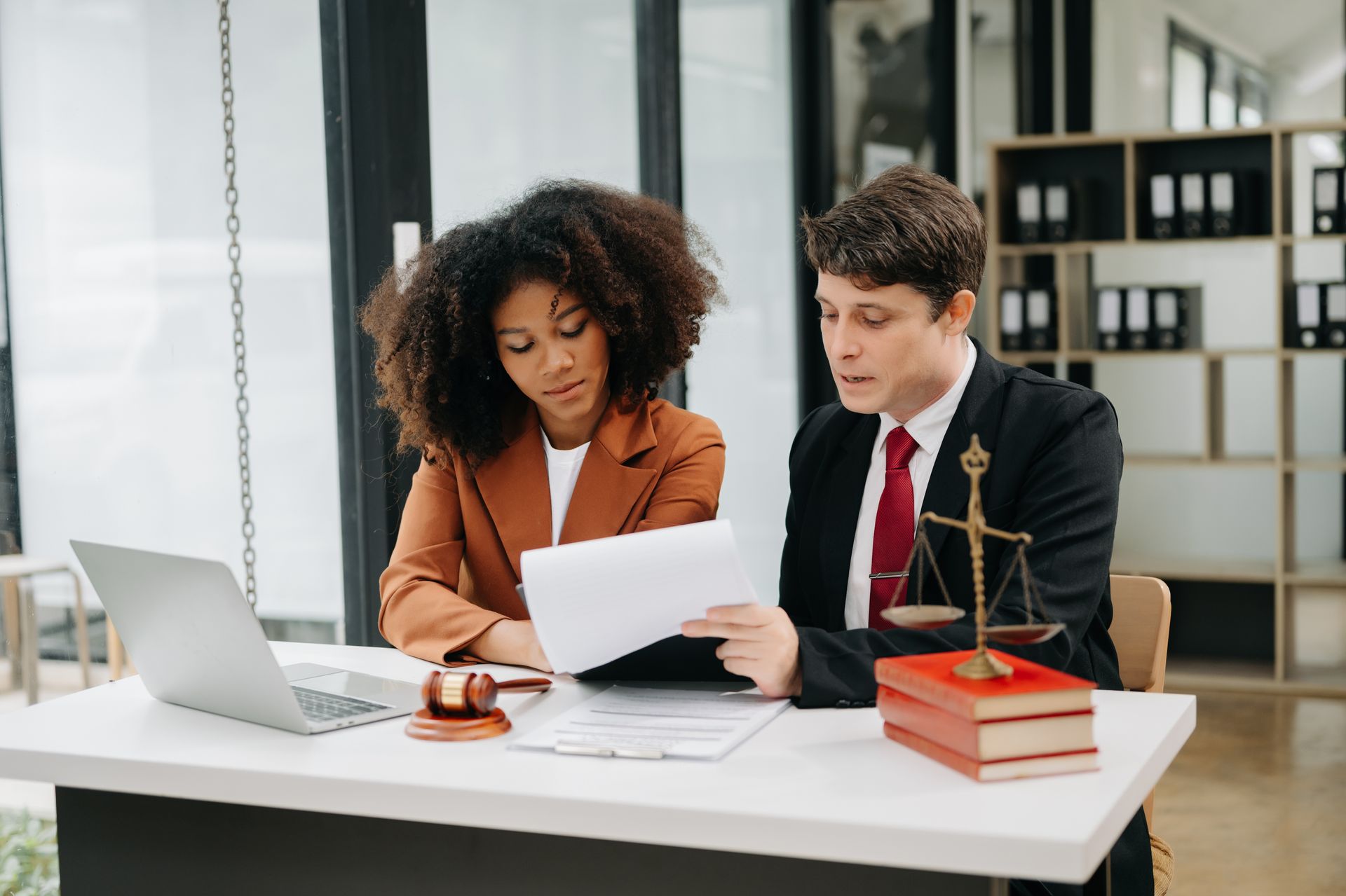 Two professionals reviewing documents at a desk with a laptop and books in a bright office
