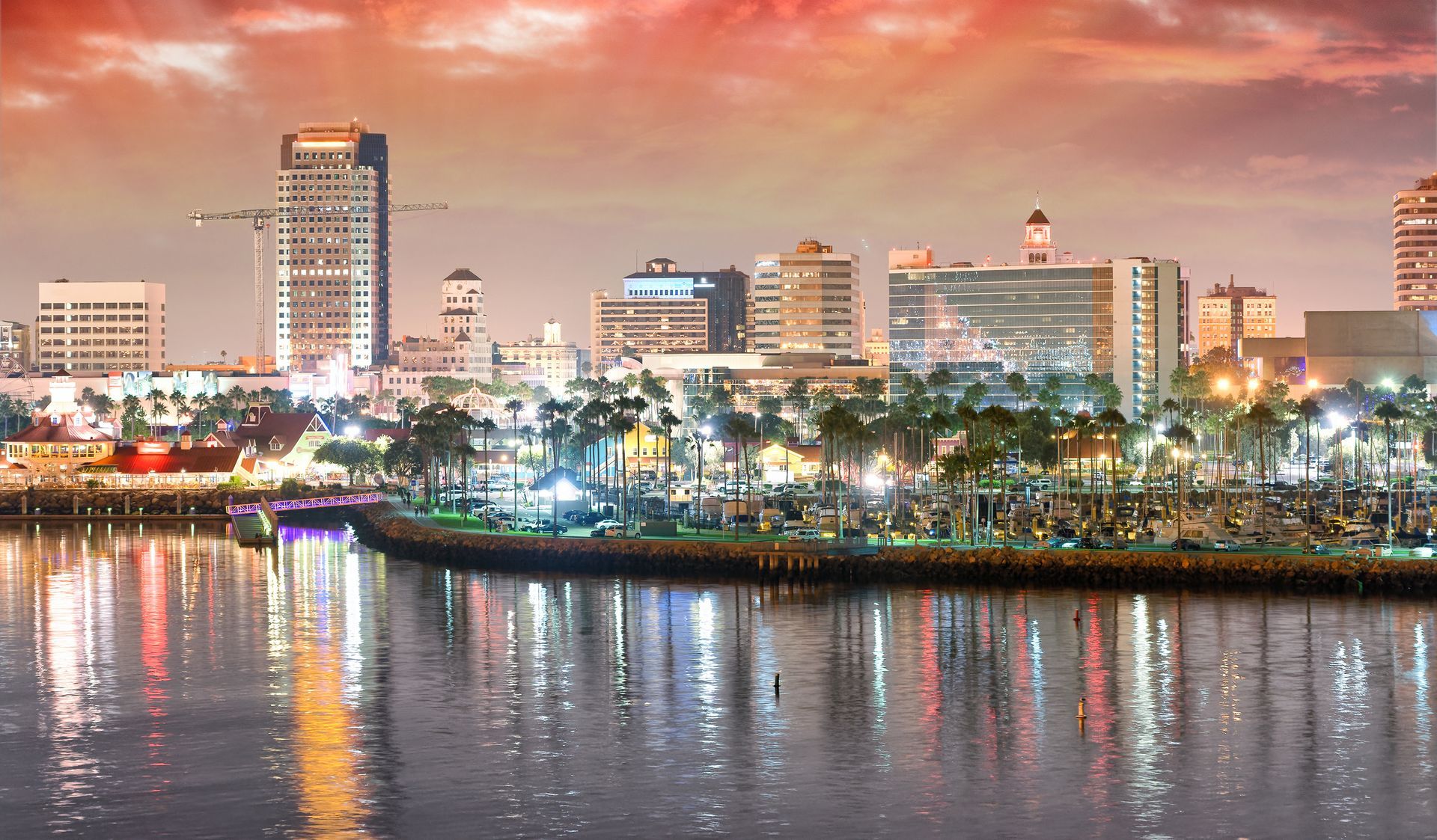 City skyline at sunset reflected on calm water, with colorful lights and pink-orange clouds.