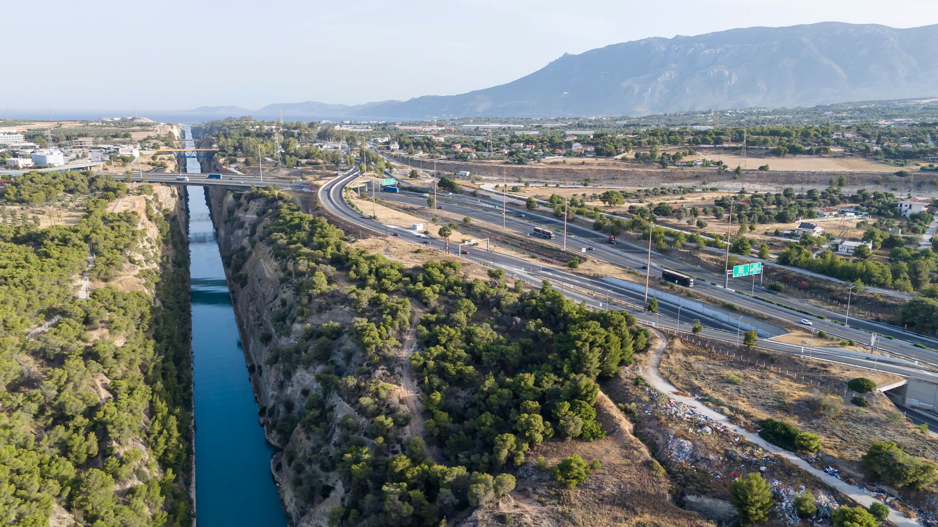 Aerial view of a highway beside a narrow canal, with green rocky hills and mountains in the distance.
