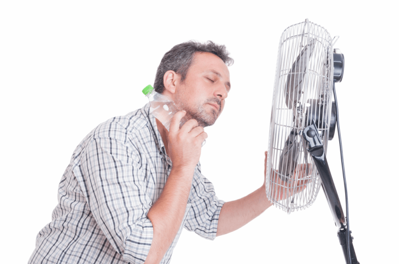 A man is drinking water from a bottle while standing in front of a fan.