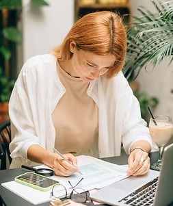Woman writing on papers at a desk with a laptop, glasses, and coffee in a bright office setting