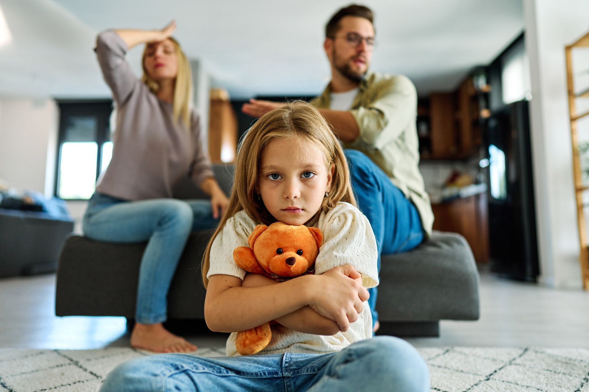 Child hugging teddy bear on couch while two adults argue in the background
