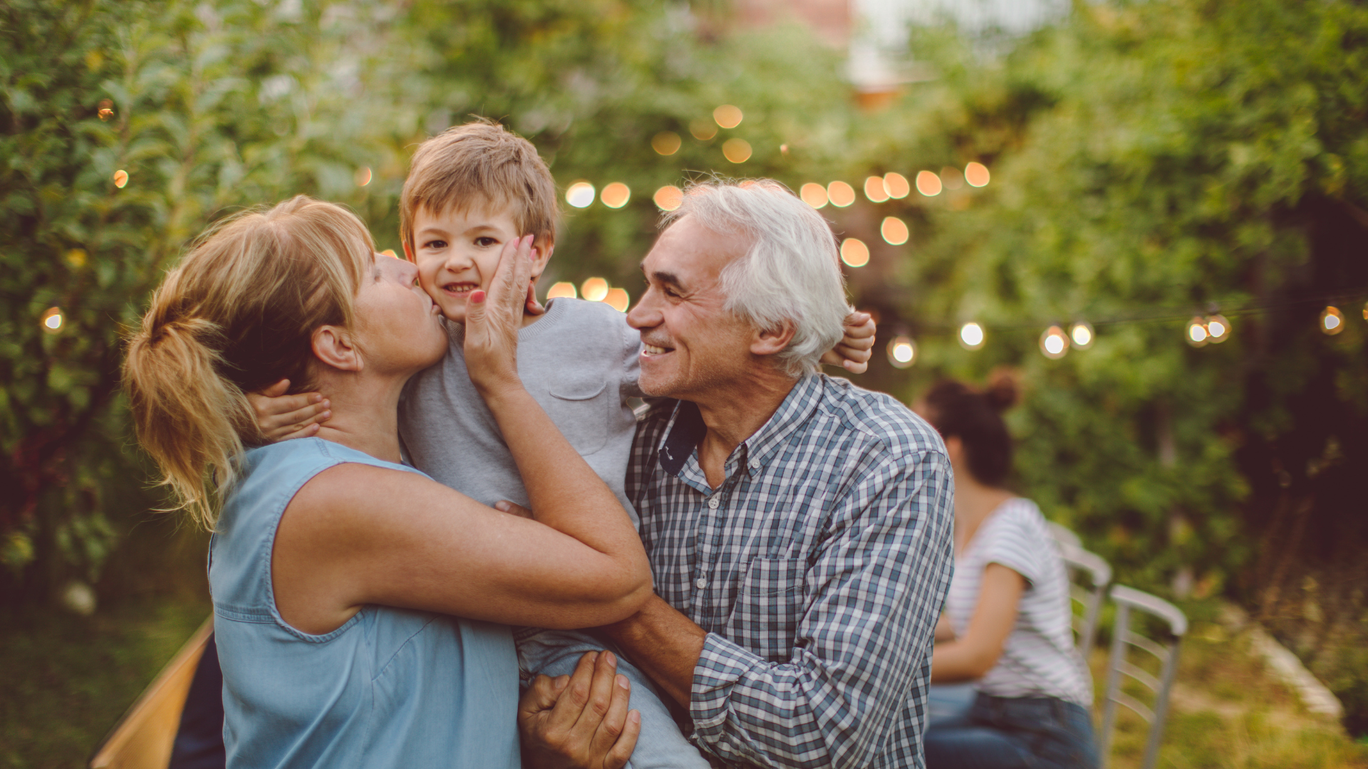 Family laughing together outdoors in a sunny garden, with a child between two adults.