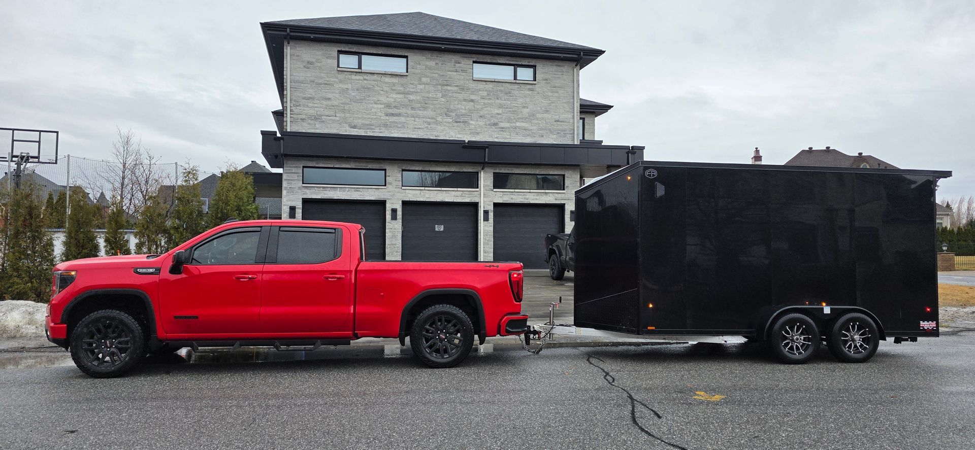 Une camionnette rouge tractant une remorque noire fermée est garée devant une maison dans une rue mouillée.