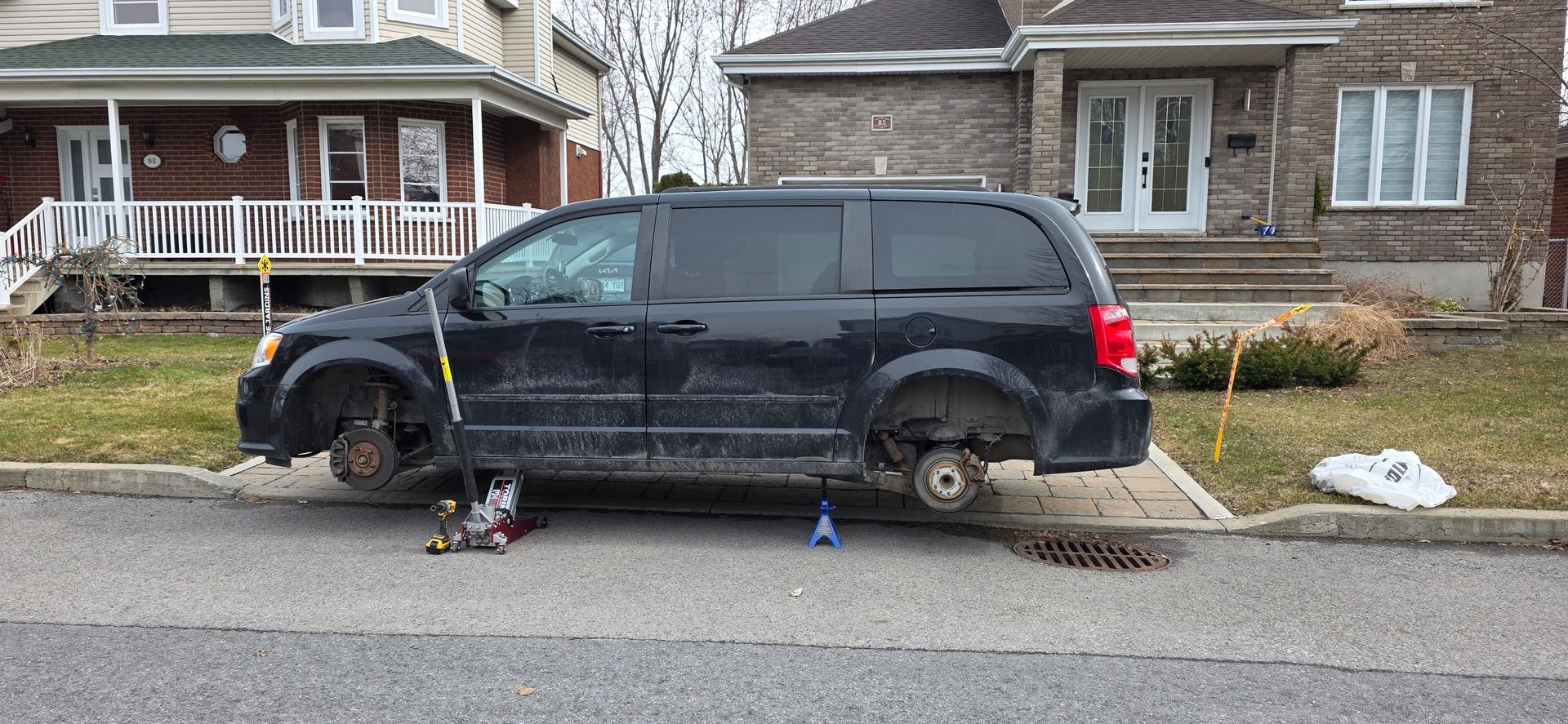 Minivan noir sans roue avant, surélevé par un cric dans une rue résidentielle, devant des maisons.