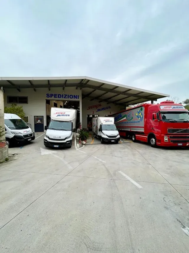 Quattro furgoni per le consegne parcheggiati all'esterno di un magazzino di spedizione sotto una tettoia metallica.