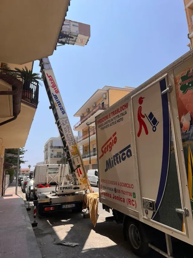 Un camion per traslochi e un'autoscala operano in una strada stretta tra gli edifici, sotto un cielo azzurro e limpido.