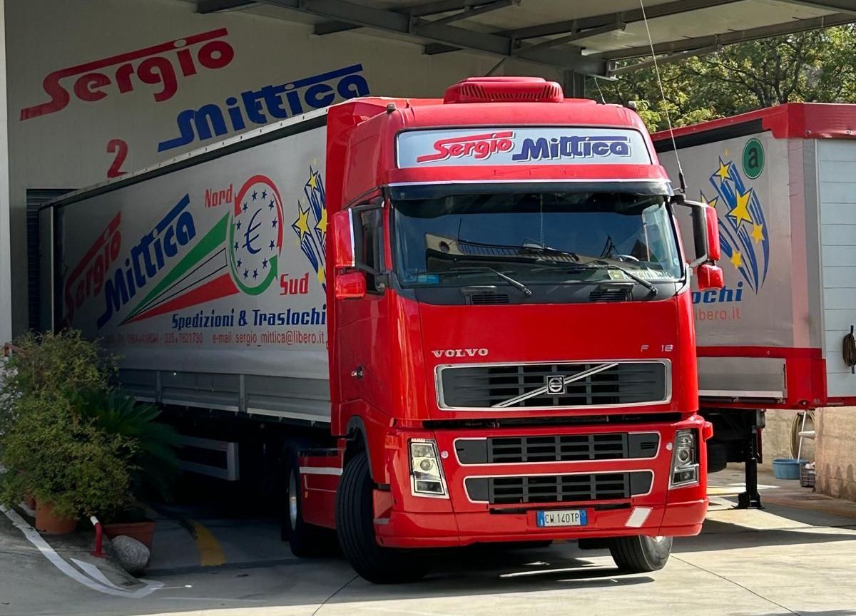 Camion Sergio rosso parcheggiato nell'area di carico di un magazzino, vista frontale con rimorchio bianco e cabina rossa.