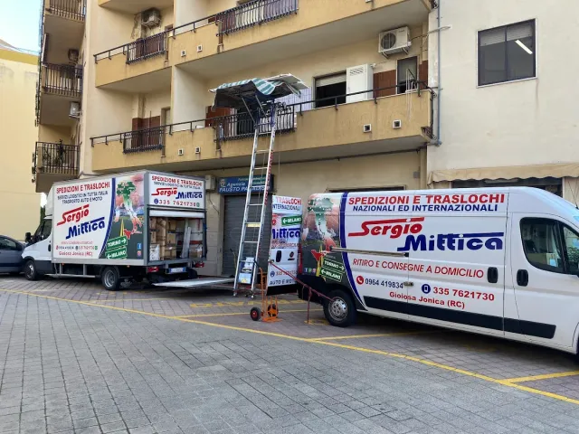 Due camion per traslochi parcheggiati vicino a un condominio, con una scala che raggiunge un balcone al piano superiore.
