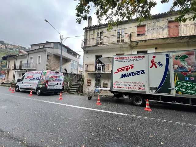 Due furgoni bianchi di servizio parcheggiati su una strada bagnata accanto a vecchi edifici, con coni arancioni lungo la carreggiata.
