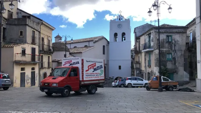 Un camioncino rosso per le consegne in una piazza lastricata di ciottoli, tra antichi edifici e il campanile di una chiesa, sotto un cielo nuvoloso.