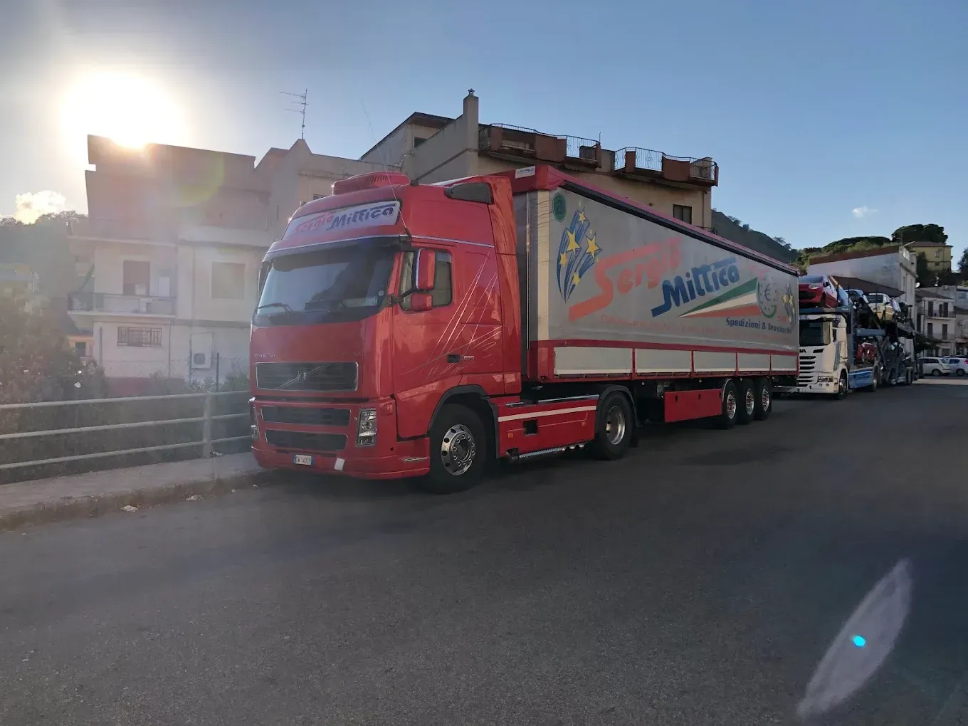 Camion Sergio rosso parcheggiato nell'area di carico di un magazzino, vista frontale con rimorchio bianco e cabina rossa.