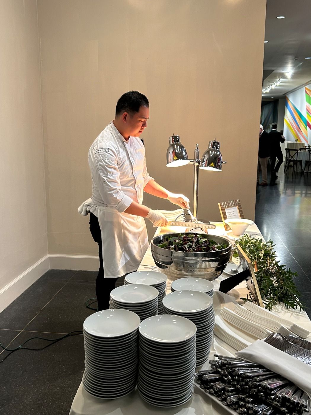 Chef serving food at a buffet table with stacks of white plates, in a well-lit modern setting.