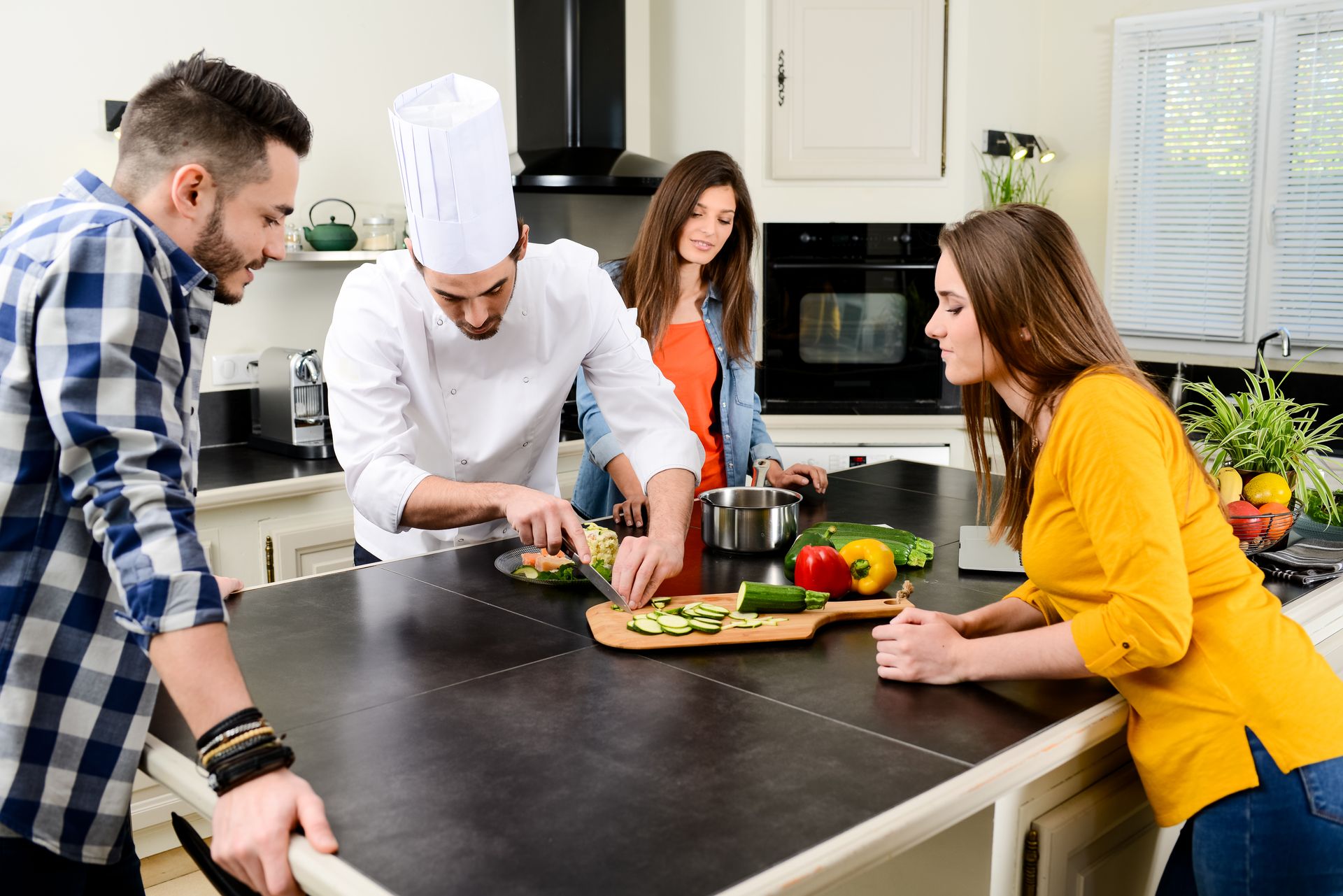 A chef prepares food on a kitchen island with three onlookers.