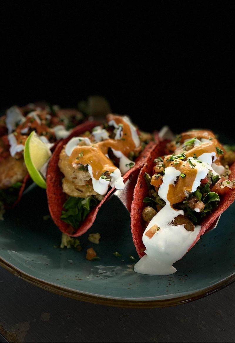 Three tacos on a blue plate, red shells filled with toppings and sauce, lime slice.