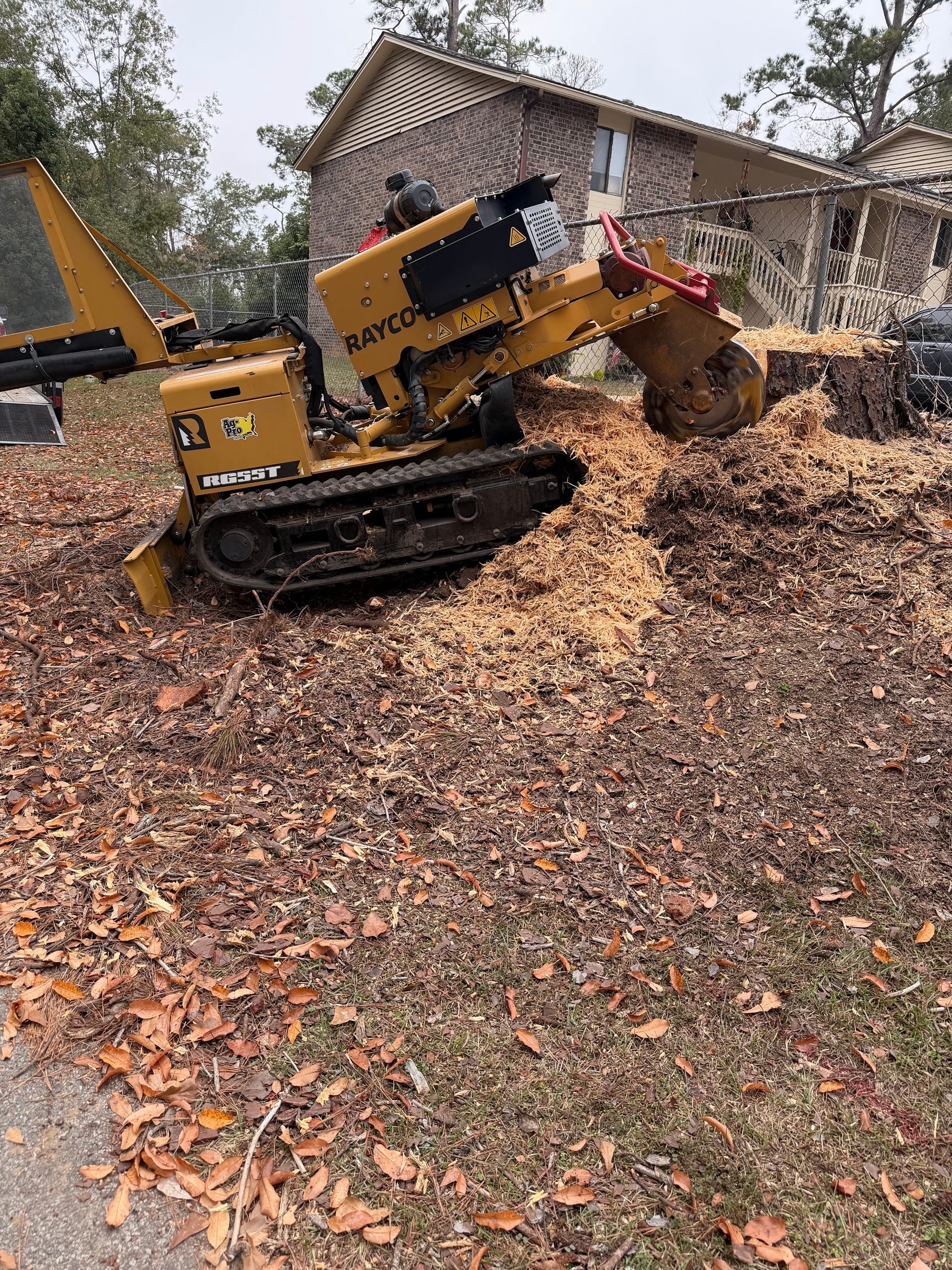 Yellow tracked skid steer lifting leaves in a yard beside a house, with a worker inside the cab.