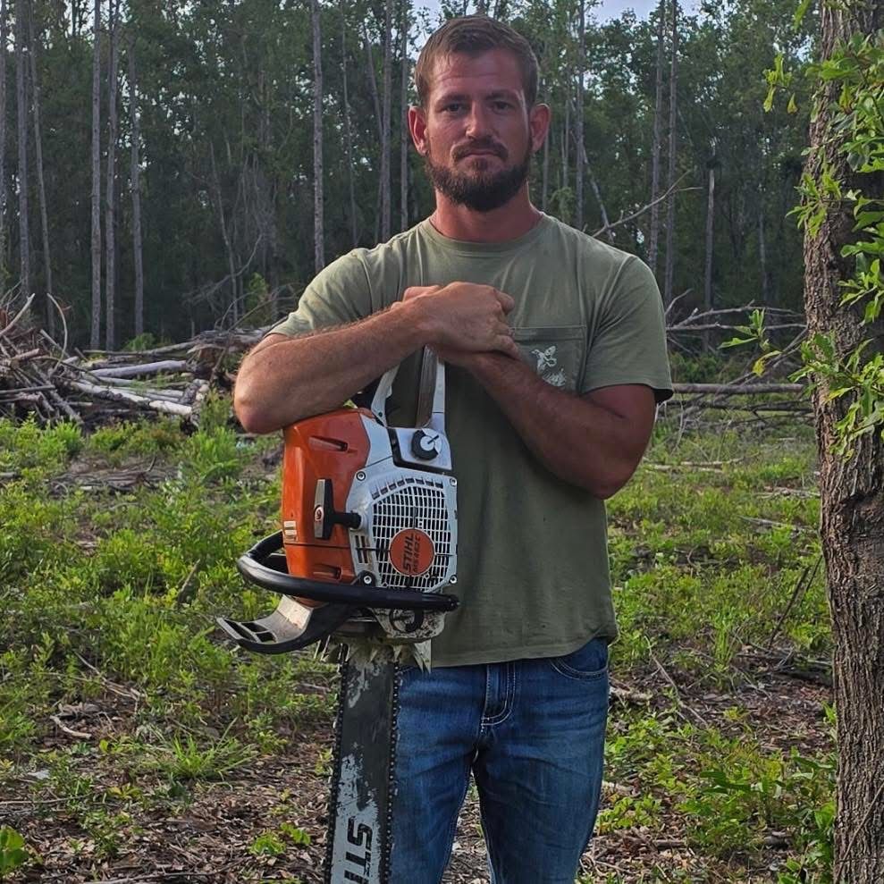 Man standing in a forest clearing holding an orange chainsaw, with cut logs and trees behind him.