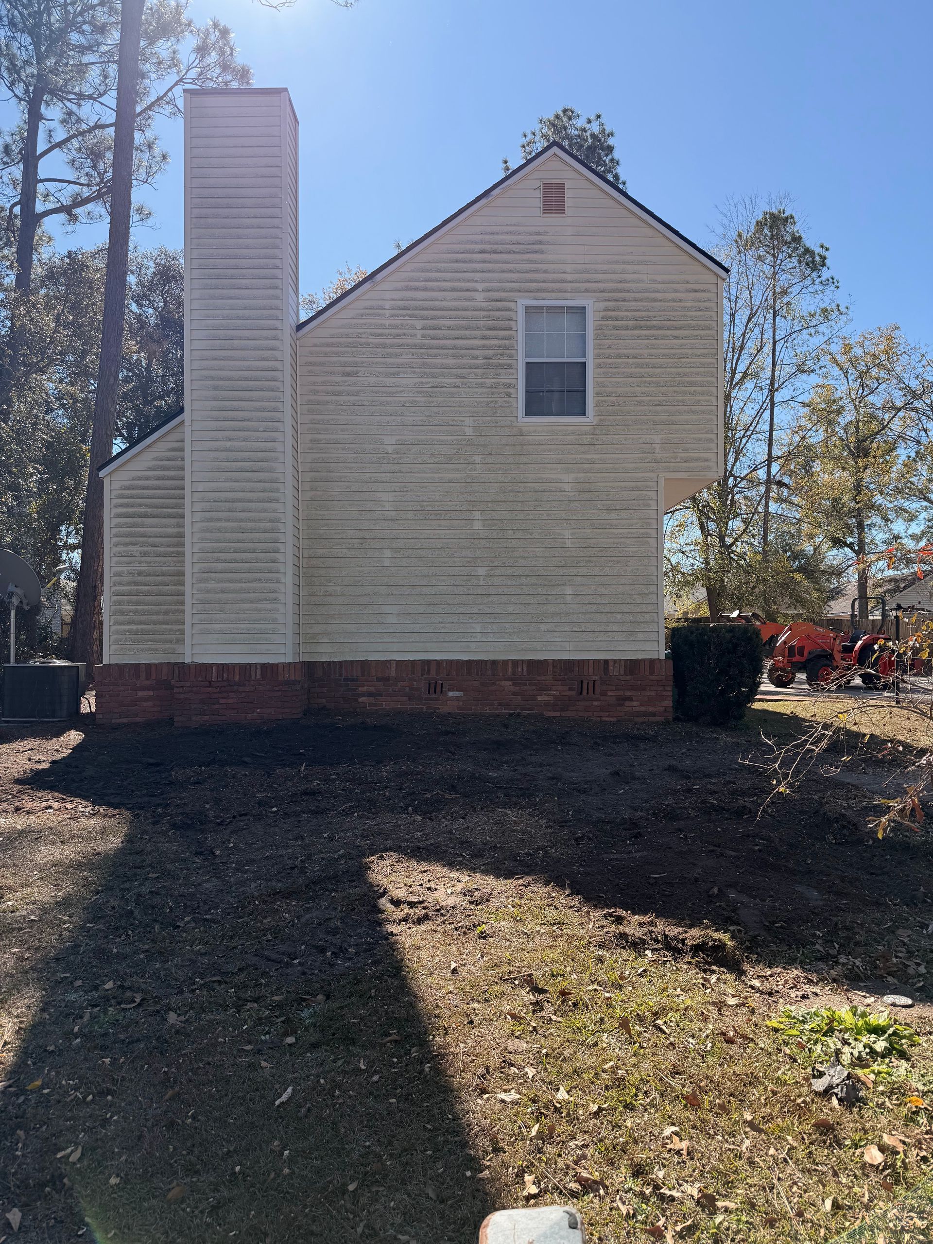 Side view of a beige house with white chimney, brick foundation, and a small upper window in a yard