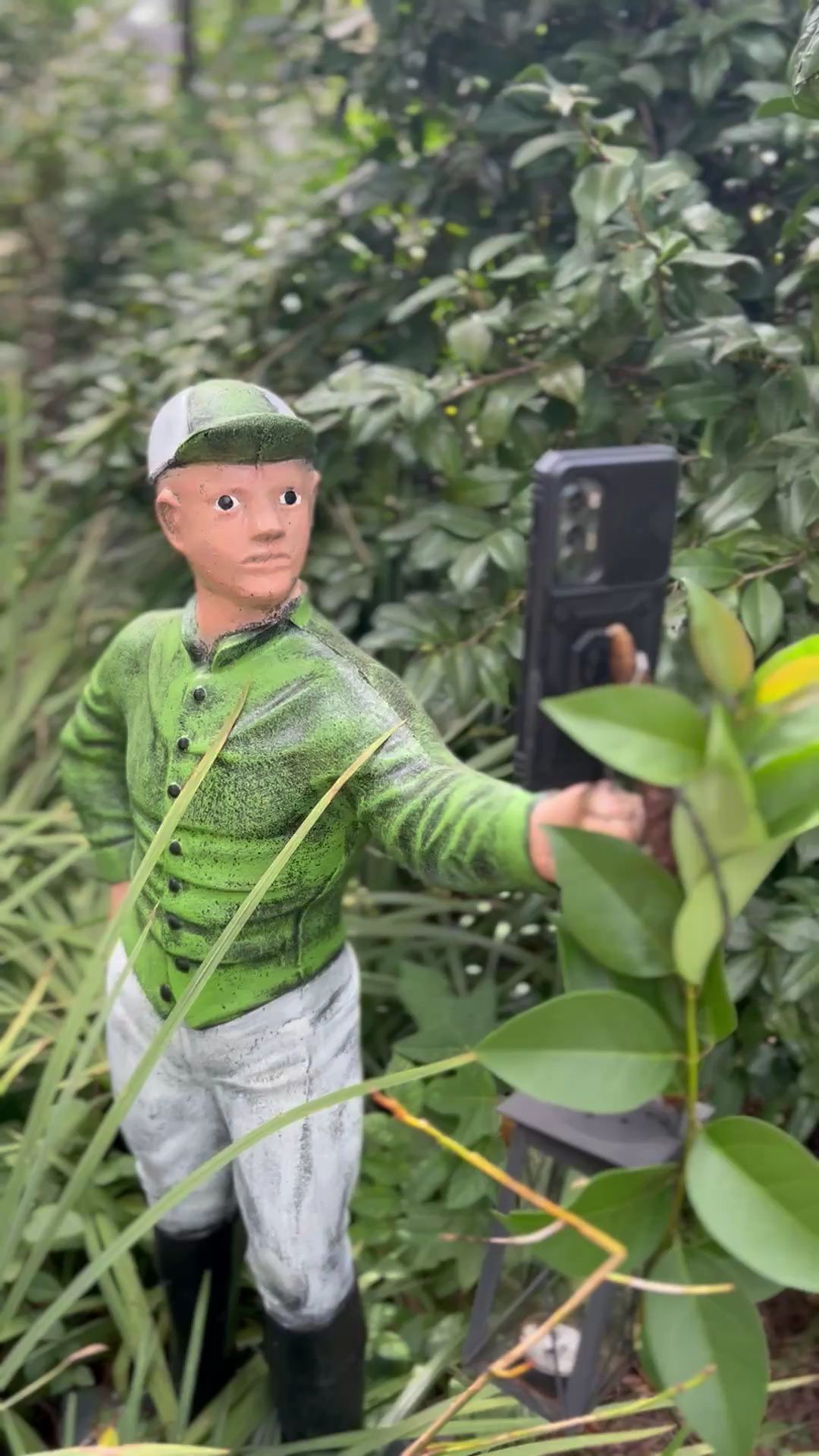 Person in green shirt and cap taking a selfie in a leafy outdoor setting with a smartphone on a tripod