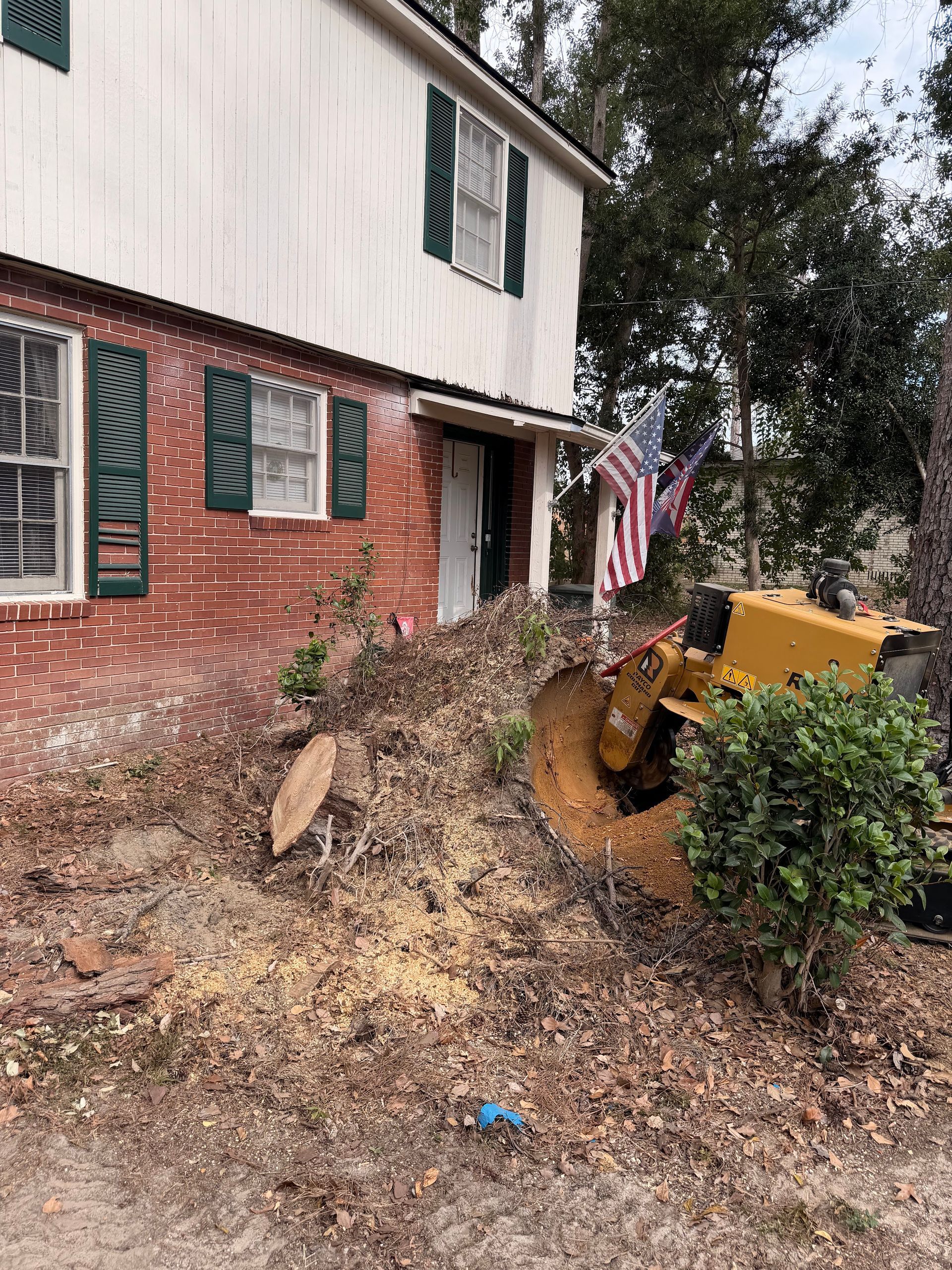 Yellow construction vehicle working on a dirt slope beside a brick house and shrubbery.