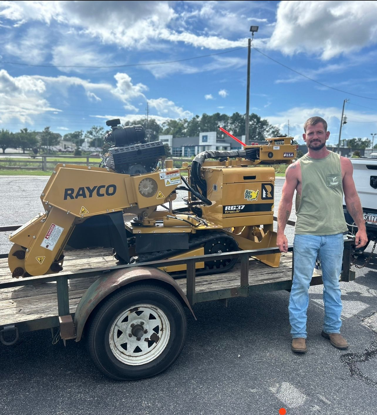 Man standing beside a yellow Rayco machine on a trailer in a parking lot under a cloudy sky
