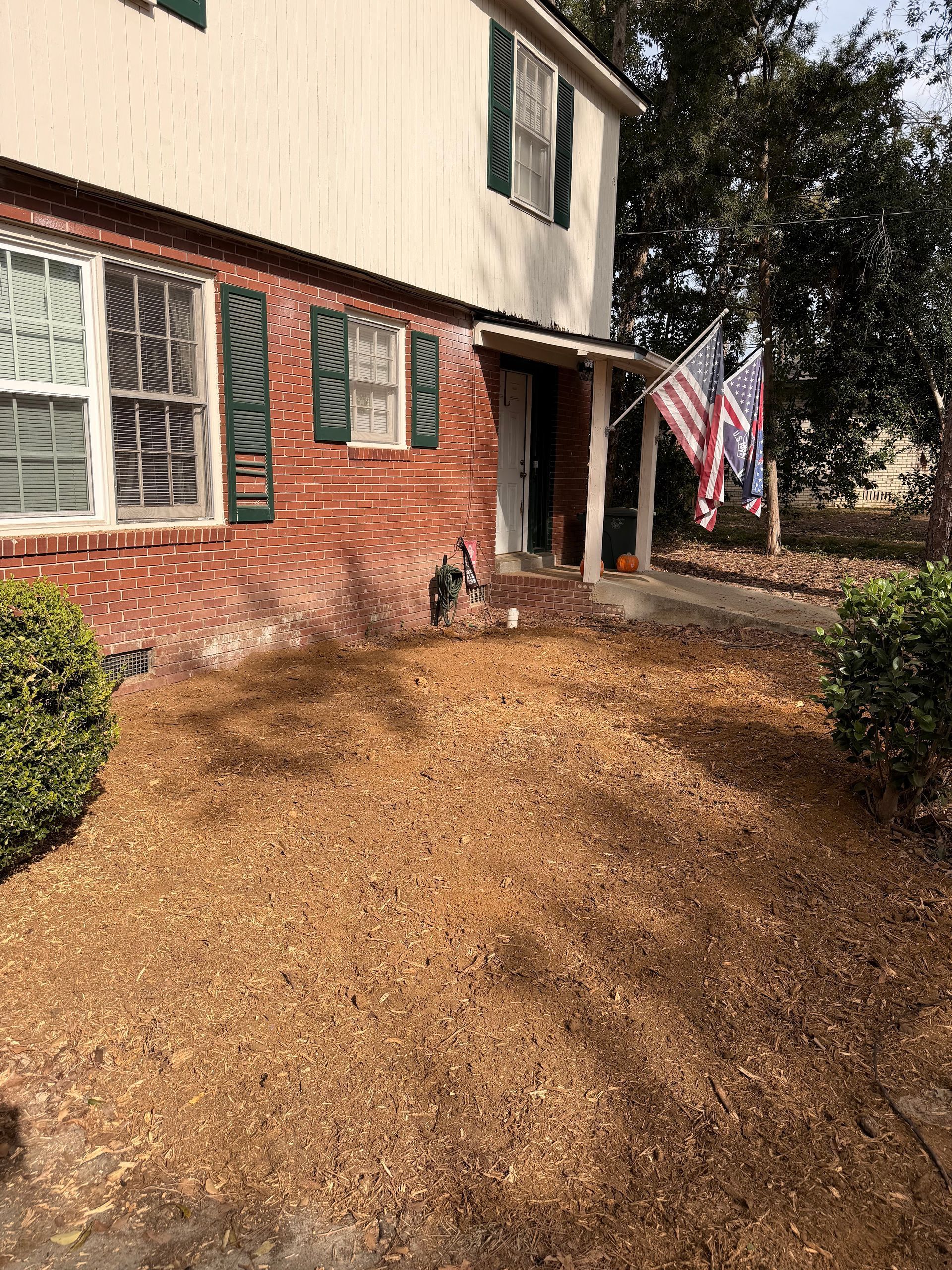 Brick house with green shutters, front porch, and an American flag beside a leaf-covered yard.