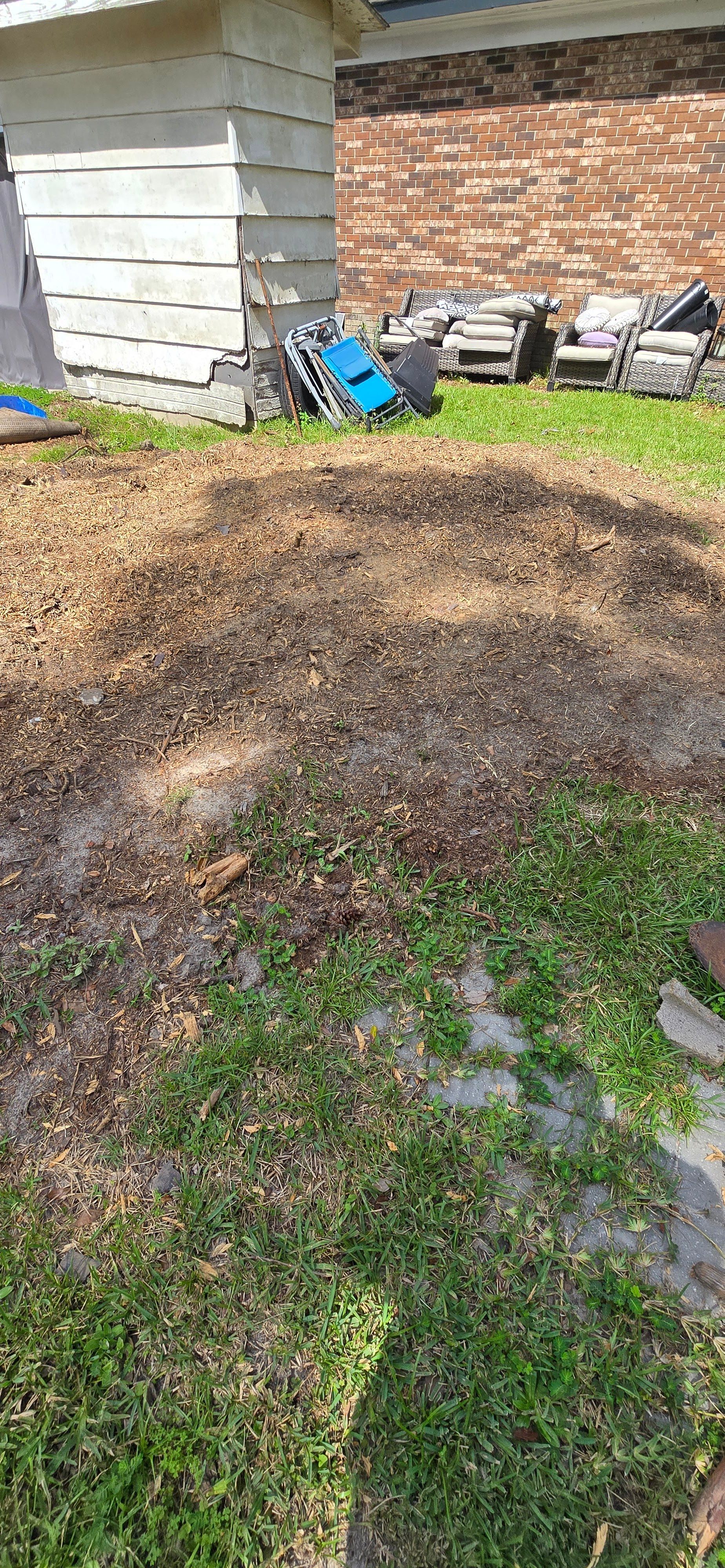 Mulch pile in a backyard beside a house, with lawn chairs and tools against a brick wall.