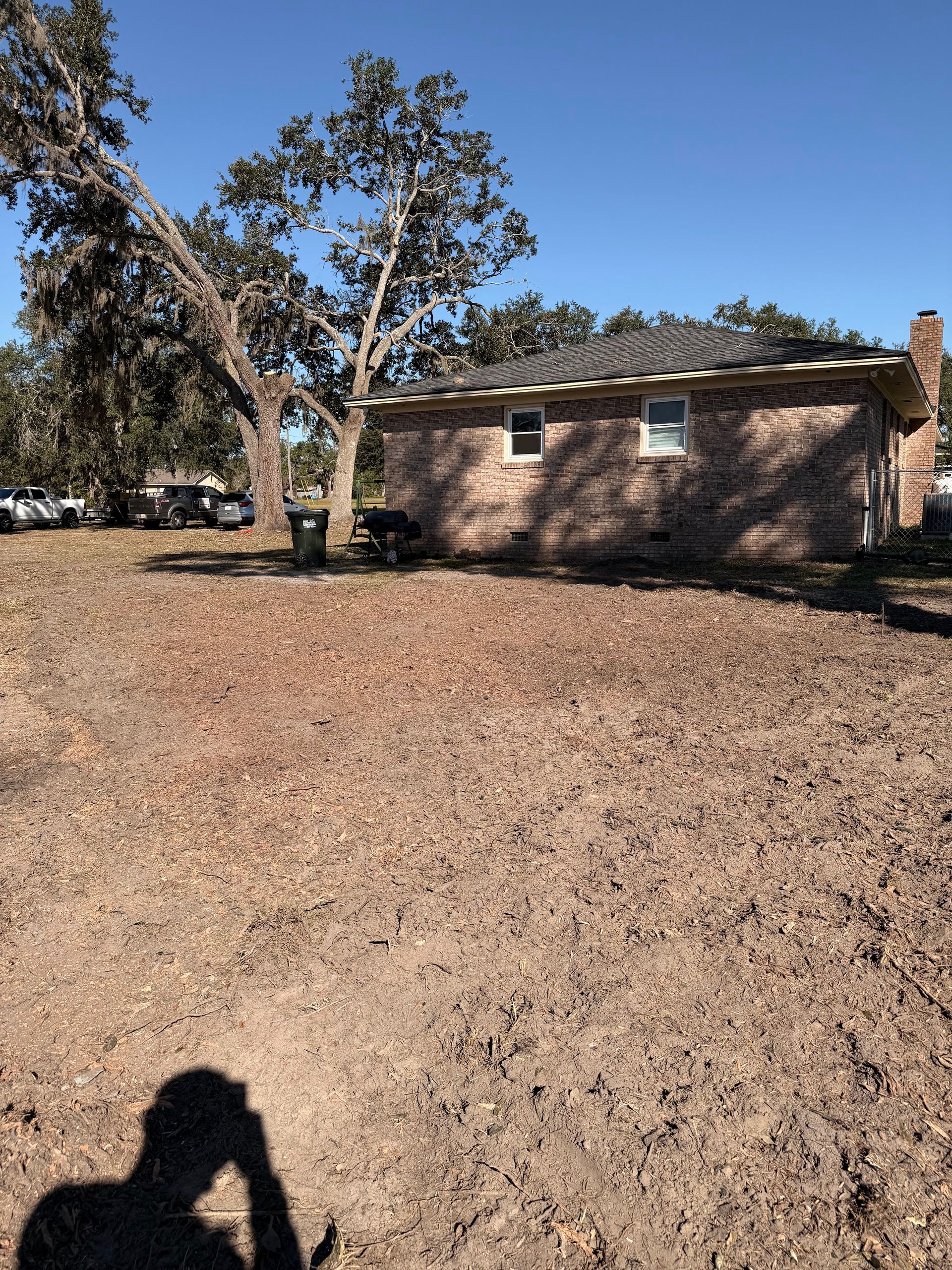Brick house behind a bare yard with large trees under a clear blue sky