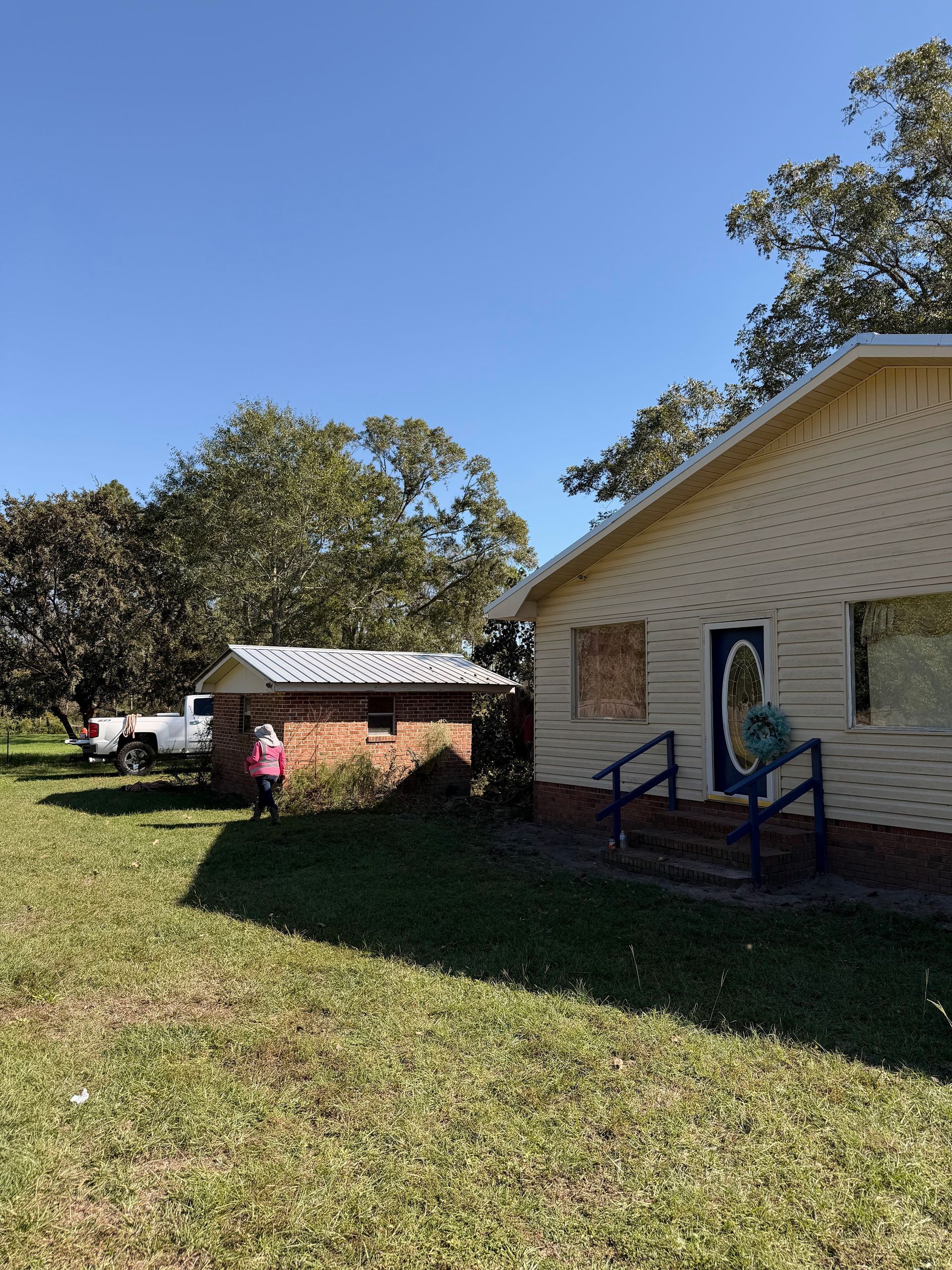 Two people near a beige house and small outbuilding on a sunny grassy yard with blue sky